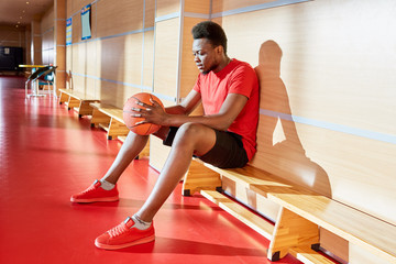 Attractive black basketball player holding ball and relaxing on bench during training in gym. 