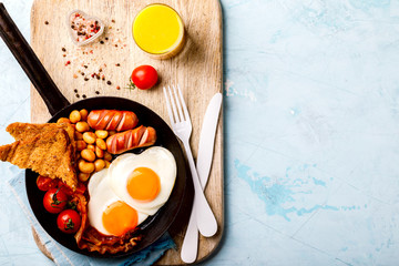 Traditional English Breakfast in the Frying Pan Eggs Heart shape. Valentine's Day. Festive Food on the Blue Background.Sausages, Bacon, Beans,Toasts,Coffe and Orange juice Top View Copy space for Text