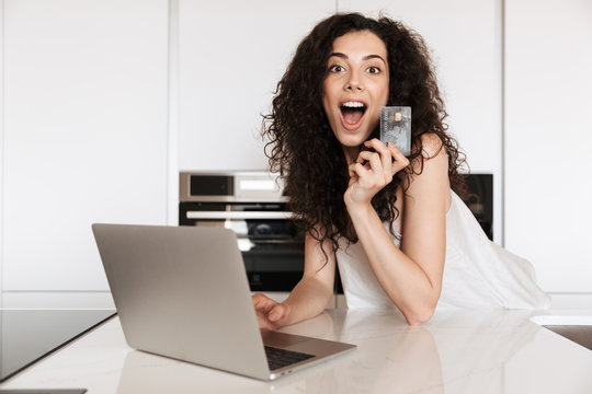 Picture of excited young woman 20s with curly brown hair wearing silk leisure clothing shouting in happiness, and shopping online with laptop and credit card while leaning on kitchen table