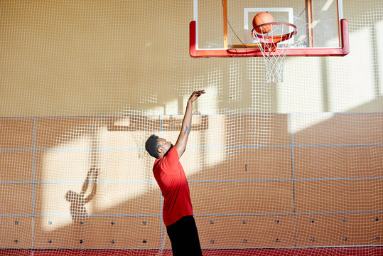 Side view of black athlete throwing ball in basket while playing basketball on gym court. 