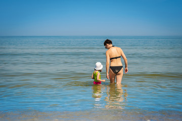 mom and little daughter in a green swimsuit are walking in the water in the sea in the summer