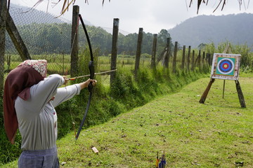 Young woman is aiming in archery  practice n the field with a target in front of her.