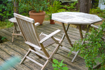 A set of wooden and lightly weathered patio furniture on a terrace made of wooden planks surrounded by green plants