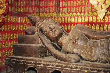 Reclining Buddha statue in a red chapel with many small Buddha statues at the background in Wat Xieng Thong temple in Luang Prabang, Laos.