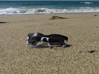 Fototapeta na wymiar Chic women’s sunglasses with sparkling reflection of the sun in one lens, isolated on a sandy beach with ocean waves in background.