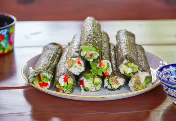 Long sushi rolls with rice, nori and vegetables lying on ceramic plate standing on wooden table with kitchenware with traditional rustic ornaments