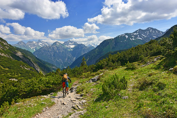 Fototapeta na wymiar A cyclist is riding a mountain bike downhill through lush-green nature. Rocky mountains and blue sky with clouds in the background. Tirol, Austria