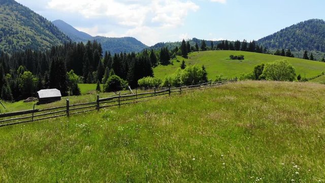 Panoramic view of idyllic mountain scenery with fresh green meadows in bloom on a beautiful sunny day in springtime. Aerial 4K view. Low air flight over mountain rural farm and meadow at sunny day