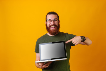 Happy smiling cheerful young bearded hipster man wearing eyeglasses and pointing at empty screen of...