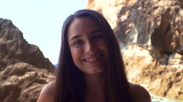 Close Up Portrait Of Beautiful Brunette Woman Turning Head At The Camera And Smiling. Rocky Sea Shore With Big Cliffs And Caves On Background