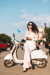 Fototapeta na wymiar Beautiful smiling lady with dark curly hair in white costume and sunglasses sitting on white moped and happily looking in camera on street with city view on background