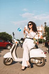 Fototapeta na wymiar Young smiling woman with dark curly hair in white costume and sunglasses sitting on white moped and happily taking photos on cellphone on street with city view on background