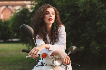 Fototapeta na wymiar Beautiful lady with dark curly hair in white costume sitting while leaning on white moped and thoughtfully looking aside on street