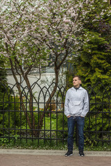 Fototapeta na wymiar Парень в белой куртке стоит под деревом в парке и ждут девушку