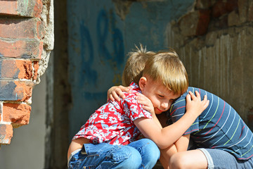 Obraz na płótnie Canvas Two boys, brothers orphans were left without housing and sit near the ruins of the building in an embrace with each other and cry and suffer in the loss. Photo production.