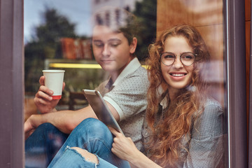 Couple of young students drinking coffee and using a digital tablet while sitting on a window sill at a college campus during a break.