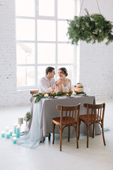 Bride and groom posing at the decorated banquet table in the white hall. Hand in hands. Enjoy a moment of happiness and love. A series of photos in my portfolio. Wedding flowers on a table