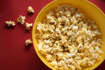 Tasty caramel popcorn in a yellow plate on the red background and next to the scattered pop corn, top view