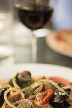 Vertical image: pasta with sea food and white sauce is on a plate in a restaurant. Background: glass of red wine, part of pizza and part of a human figure in blue clothes.