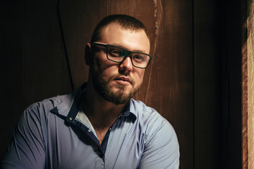 Fototapeta premium brutal bearded man with a tattoo on his arm, portrait of a man in dramatic light against a brown wooden wall, bearded male with tattoo dressed in a shirt, man wearing spectacles looks in the side