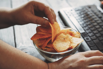 Unhealthy snack at workplace. Hands of woman working at computer and taking chips from the bowl....