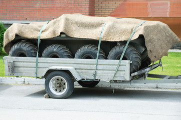 The trailer is loaded with automobile tires and covered with a cloth. It is in the street in the summer.