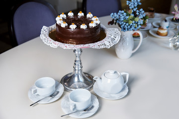 The chocolate cake on the stand stands on a table with flowers, various desserts, cups and a teapot.