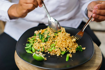 Men's hands are eating spoon mash a stir fried Mama or Thai phat  fried noodles with sliced lemon and vegetables in black dish, On the Oak wooden table background.