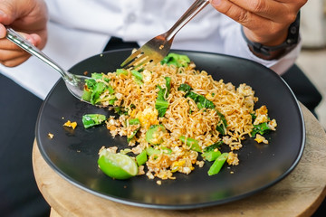 Men's hands are eating spoon mash a stir fried Mama or Thai phat  fried noodles with sliced lemon and vegetables in black dish, On the Oak wooden table background.