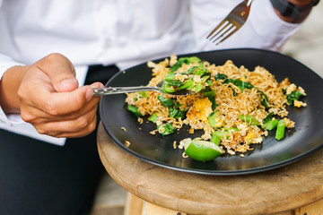Men's hands are eating spoon mash a stir fried Mama or Thai phat  fried noodles with sliced lemon and vegetables in black dish, On the Oak wooden table background.