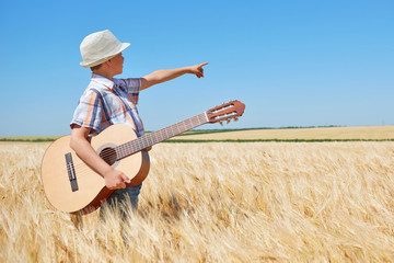 child boy with guitar is in the yellow wheat field, bright sun, summer landscape