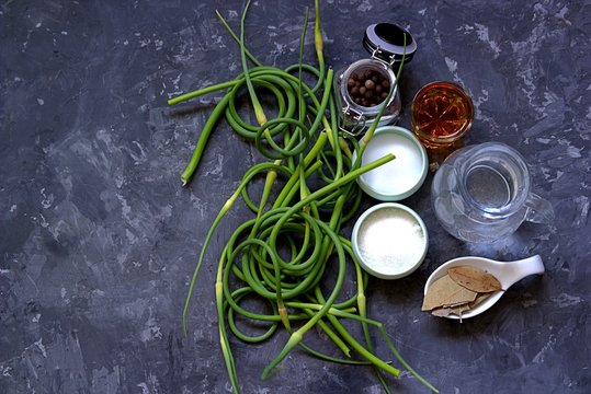 Ingredients for marinating fresh young garlic arrows on a dark gray concrete background. Fresh garlic hands, salt, sugar, vinegar, bay leaf, water, sweet pepper.