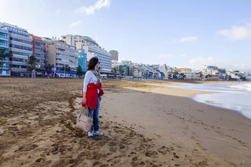 Poster Las-Palmas de Gran Canaria, Spain, on January 10, 2018. The winter sun lights the Playa de Las Canteras beach and the beautiful embankment in the distance. The attractive woman walks at a water edge © Elena Belyaeva