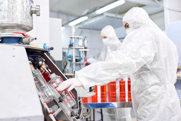 Sports nutrition production worker in protective clothing placing empty bottle into filling...