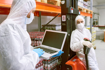 Sports nutrition production worker standing in warehouse in protective clothing and using laptop...