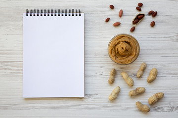 Bowl of peanut butter, peanuts and notebook on a white wooden surface, top view. From above.
