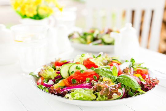 Fresh vegetarian vegetable salad with tomato, onion, cucumber, pepper, basil and lettuce. Salad on plate on white table