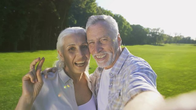 Beautiful aged senior couple making self-portrait photos on cellphone outdoors. Happy smiling married elderly wife and husband posing while making selfies on mobile phone to share in social media