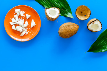 Pieces of coconut flesh on plate on blue background with coconut cut in half and palm leaves top view copy space