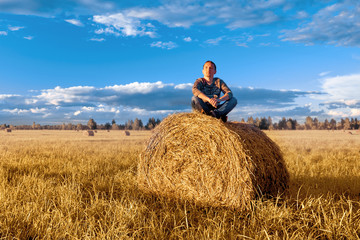 Naklejka na ściany i meble A young man sitting on a haystack, against the blue sky with clouds