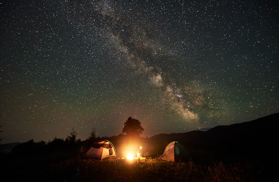 Woman camper resting at night camping in mountains under amazing night sky full of stars and milky way. Girl hiker sitting beside campfire and two tents. Concept of active lifestyle. Astrophotography
