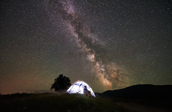 Female tourist resting at night camping in the mountains under incredible beautiful starry sky and Milky way. Woman sitting inside illuminated tent and looking at sky full of stars. Astrophotography