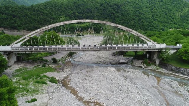 Aerial view. On the old bridge over a mountain river passes a white car. The camera moves away from the bridge and offers stunning views of the mountain gorge.