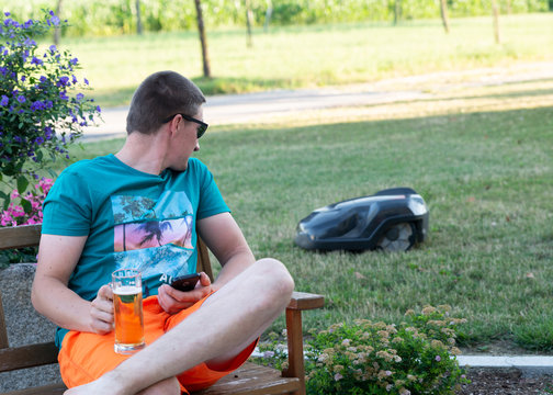 sportlicher Mann sieht dem M&auml;hroboter zu, Urlaub, Bier, Sonnenbrille, Handy, Rasenroboter