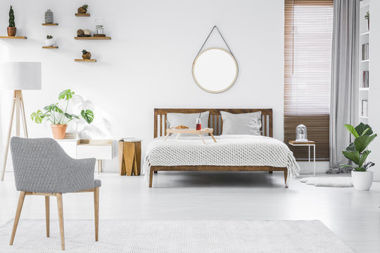 Gray, modern armchair, breakfast tray on a double bed and wooden furniture in a simple, stylish apartment room interior with white walls