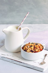 Cup of muesli with tropical fruits and white milk jug on wooden board. Concept of healthy eating in pure tones.