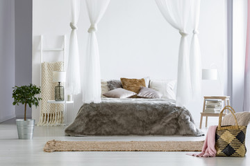 Fur coverlet placed on king-size bed with cushions and canopy in white bedroom interior with carpet, straw bag with pillow and blanket, fresh plant standing on the floor and books on small table