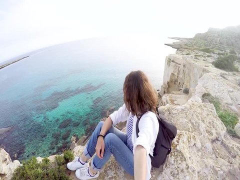 A young woman with a backpack sits on a cliff above the sea and looks at the horizon. Selfie on the background of a beautiful view.