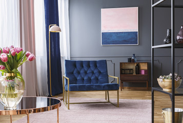 Navy blue armchair next to lamp in sophisticated apartment interior with painting and flowers. Real...