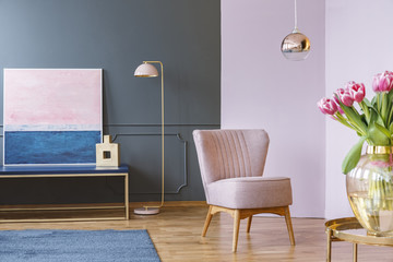 Pink tulips on table in pastel living room interior with armchair and lamp near painting. Real photo